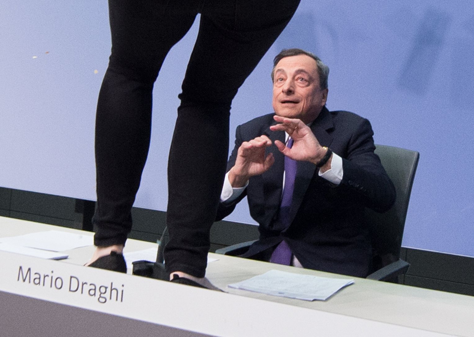 Eine Aktivistin stört am 15.04.2015 die Pressekonferenz der Europäischen Zentralbank in Frankfurt am Main (Hessen) und steht dabei vor dem EZB-Präsidenten Mario Draghi auf einem Tisch. Sicherheitskräfte der Bank führten die Frau ab. Näheres zu den Hintergründen der Aktion war zunächst nicht bekannt. Foto: Boris Roessler/dpa +++(c) dpa - Bildfunk+++